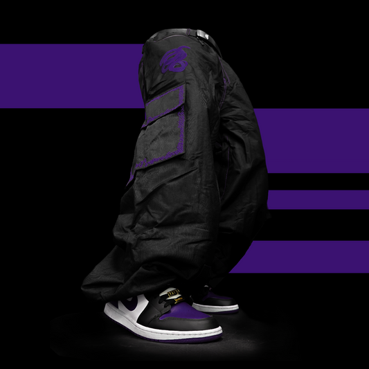 A pair of baggy snowpants (ski pants or snowboad pants) stand on a black background. they do not have a torsoe, but appear to be lifelike. the pants are black, with colorful embroidery around the pockets. purple embroidery. there are two pruple snakes weaving in and out of each other, embroidered above the right pocket.