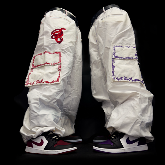 The two pairs of baggy snowpants stand facing each other. they do not have a torsoe, but appear to be lifelike. the pants are white, with colorful embroidery around the pockets. one of them has red embroidery, the other has purple embroidery. there are 2 snakes embroidered on the pant, weaving in and out of each other. 